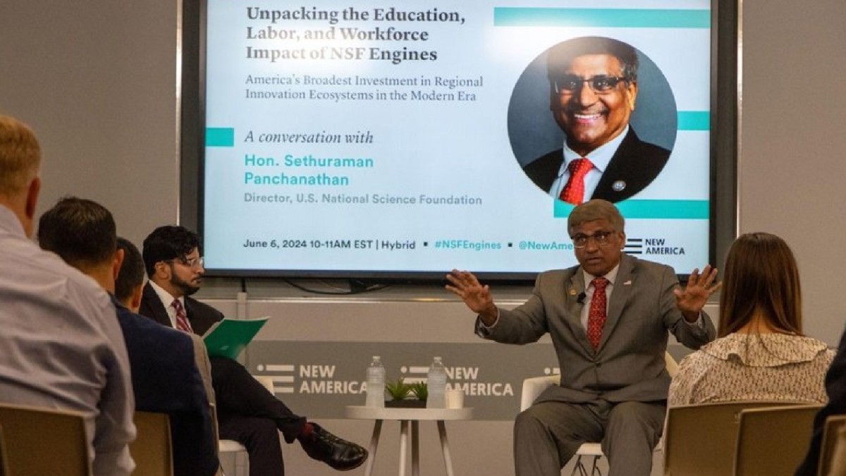 A man in a business suit speaks to students about a presentation that is projected on a screen in the background.