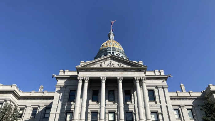 With the camera positioned beneath the Colorado State Capitol in Denver, we gaze up at its imposing marble columns, golden dome, and American and Colorado flags flying under a cloudless blue sky. 