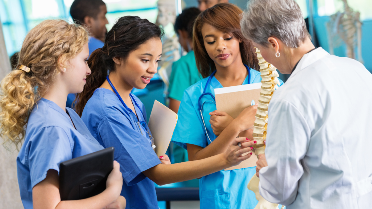 Three female nursing students hold documents and interact with their instructor and a skeleton for medical students.