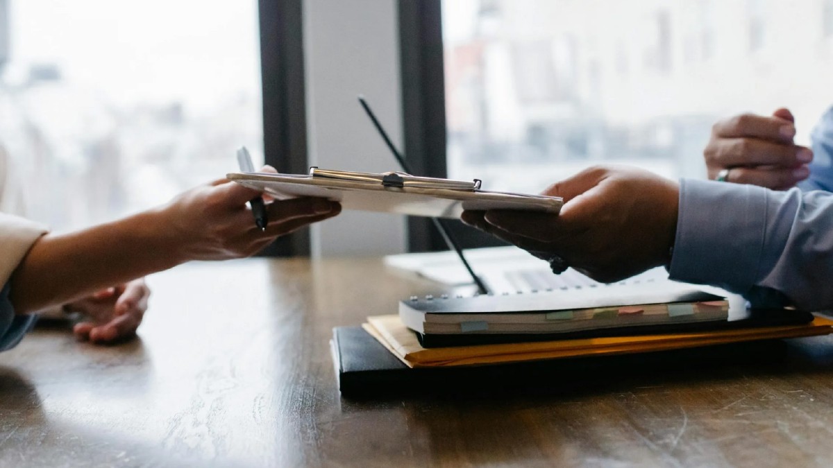 In an office setting, a male hand passes a clipboard over a desk filled with notebooks to a female hand holding a pen. 