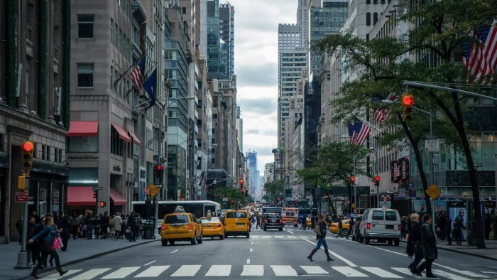 A busy Manhattan street is flanked by skyscrapers as workers cross the street behind yellow taxis and other cars. 