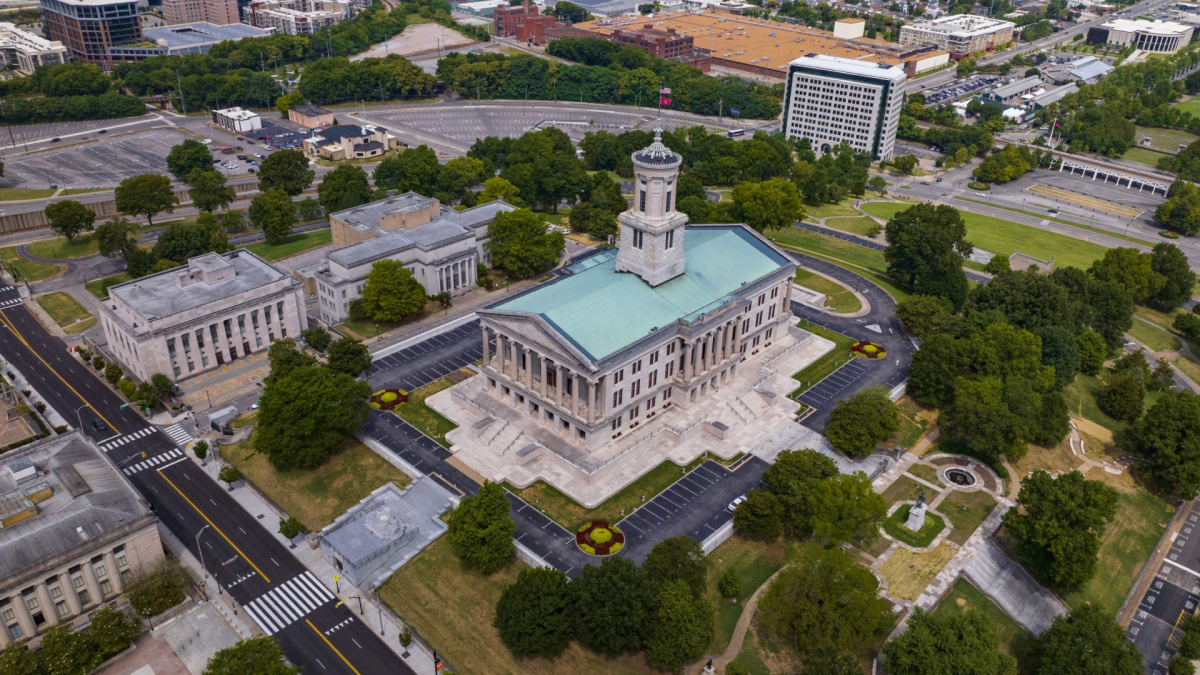 An aerial shot of the Tennessee State Capitol centers the large white building with its nickel green roof and tower jutting out surrounded by urban Nashville roads and buildings.