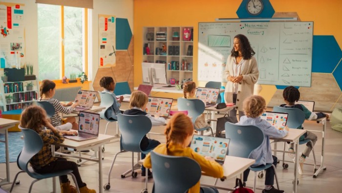 In a bright and richly decorated classroom, a young female teacher looks out at eight elementary school students who are working on laptops at their desks.