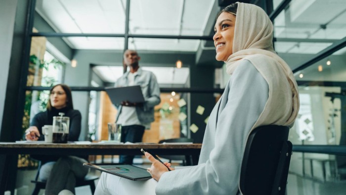 While sitting in a minimalist office space, a smiling woman in a headscarf participates in a business meeting with two engaged colleagues in the background.