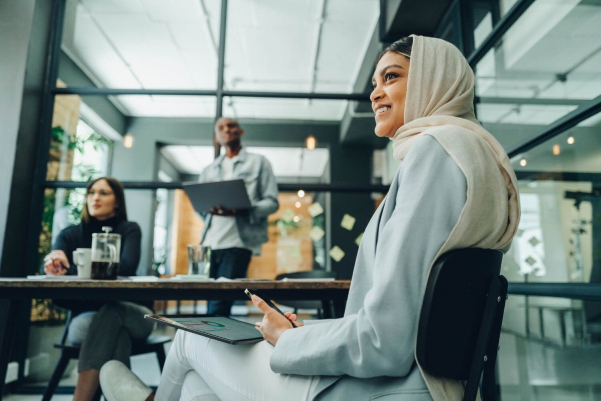 While sitting in a minimalist office space, a smiling woman in a headscarf participates in a business meeting with two engaged colleagues in the background.