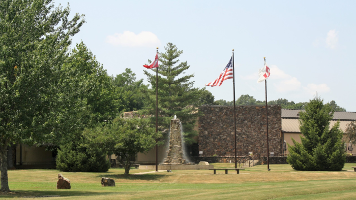 Three flags, planted in the mowed lawn of North Arkansas College, blow in the breeze before a stone water fountain that leads to a matching academic building.