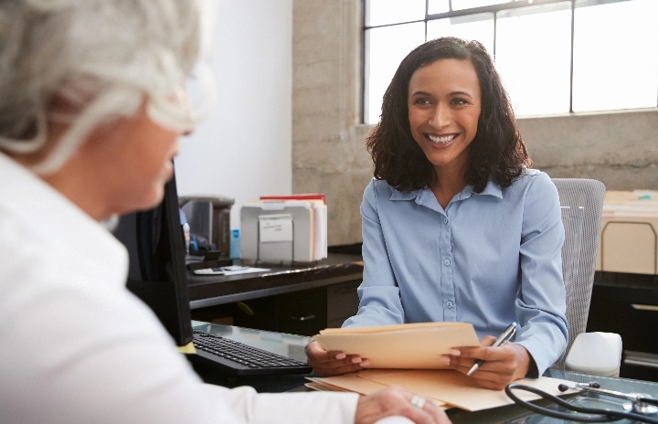 An older woman with light hair sits across a desk from a woman holding sheets of paper and smiling.