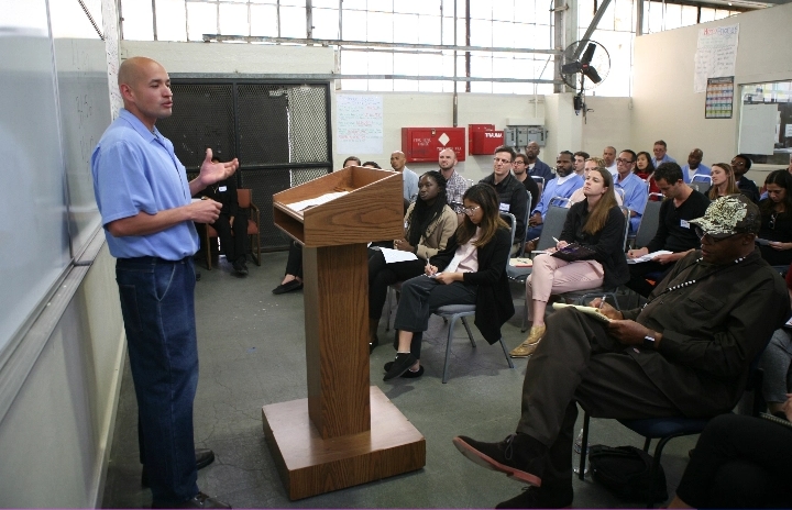 A bald gentleman in a blue collared shirt stands in front of a podium addressing a group of seated people.