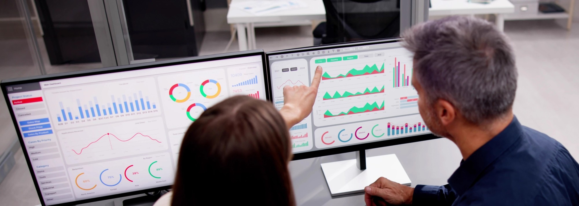 Looking over the shoulders of a man and woman focused on 2 computer monitors with many colorful graphs