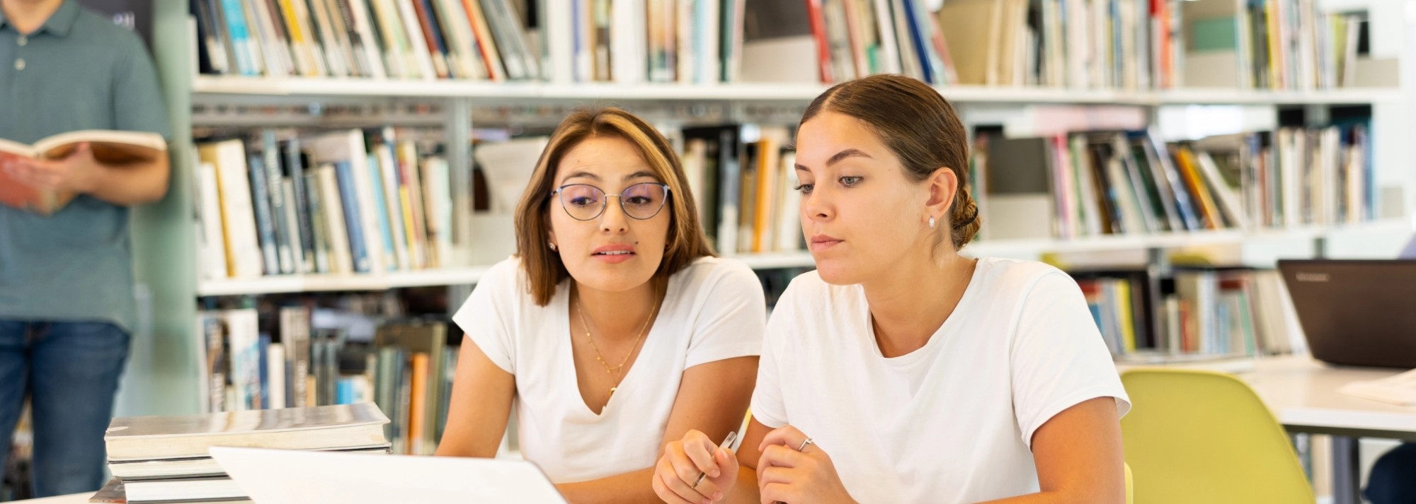 Two young women in white t-shirts focus on a laptop and various notes. Behind them is a large bookshelf.