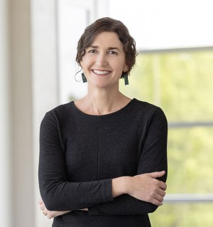 A photo of Jessa Valentine smiling with arms crossed and standing in front of a sunny window