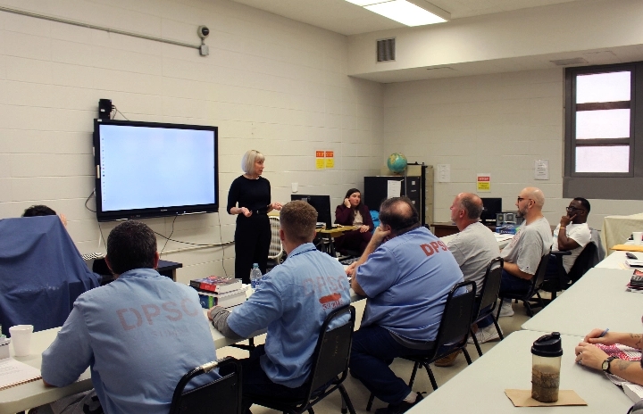 Within a cinder block room a woman with grey hair stands in front of a TV monitor addressing several justice involved men who are facing her and sitting at a long table beside one another.