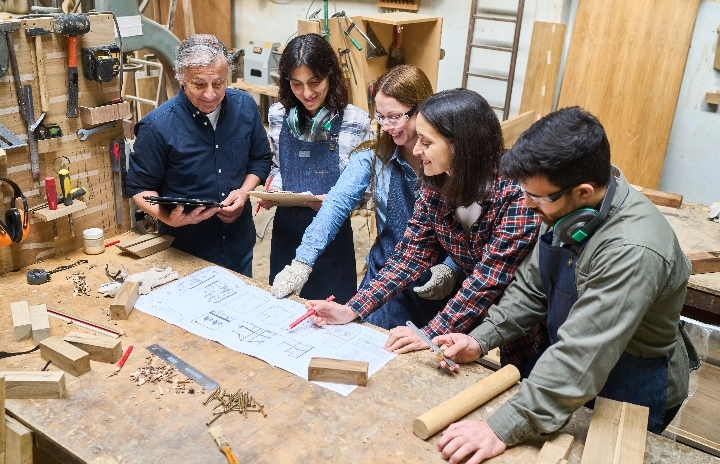 4 students and a teacher stand over a table in a wood shop looking at blueprints.