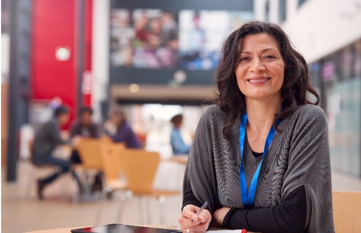 In the immediate foreground a middle aged woman holds a pen and is smiling at the camera. Behind her and out of focus are several people sitting at tables.