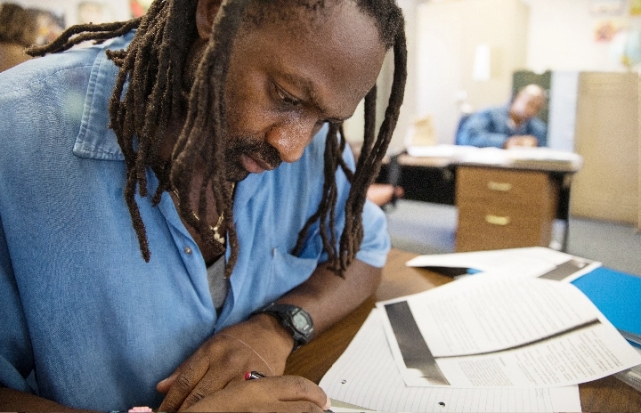 And older black man with braided hair wearing a blue collared shirt holds a pen and is focused intently at lined paper on the desk before him.