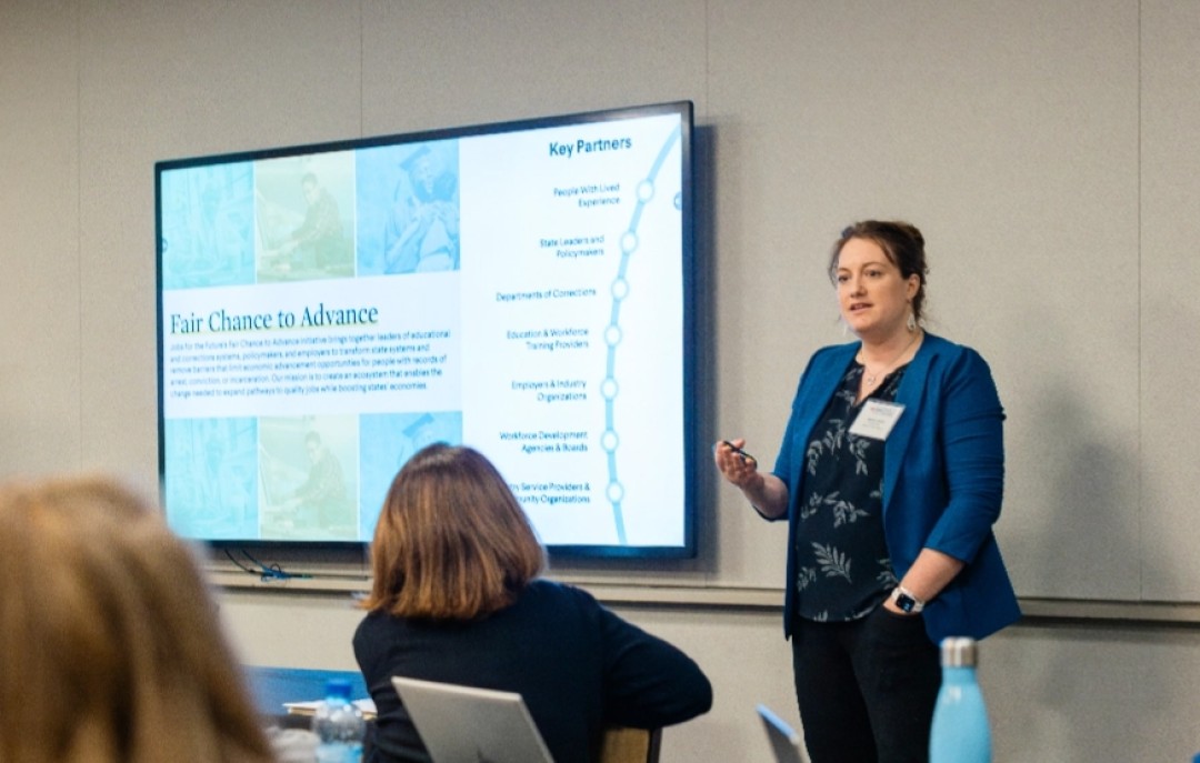 A woman dressed in a blue blazer stands beside a large screen. The screen includes the words: Fair Chance to Advance. She is addressing a seated audience.