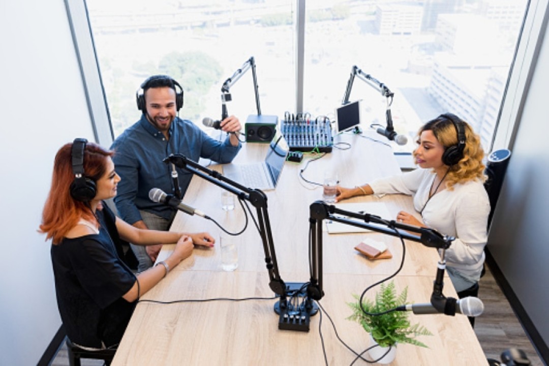 A man and a woman in headphones set across the table from a woman also in headphones. The all have microphones on arms facing them. They are engaged in conversation.