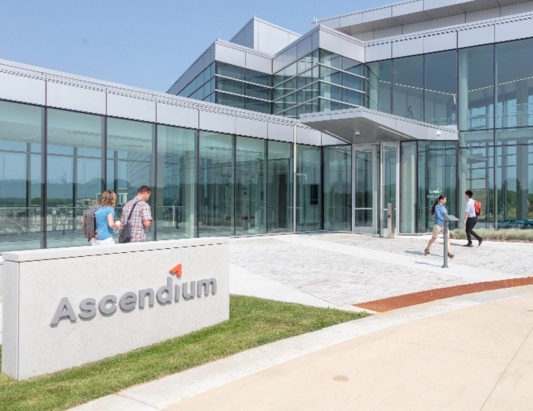 Outside on a sunny day the a sign in front of a glass walled building bears the letters spelling Ascendium. Several people are entering and exiting the building.